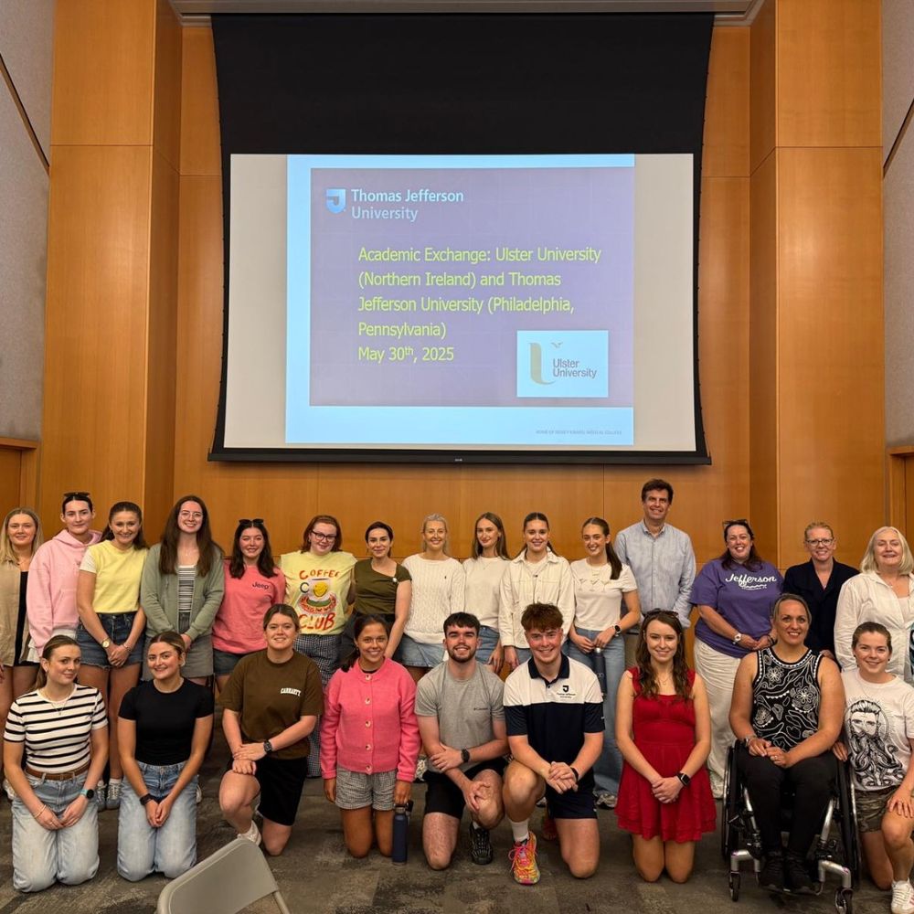 A large group of adults, including men and women, pose for a photo in a lecture hall. A screen in the background displays a presentation slide titled "Academic Exchange: Ulster University (Northern Ireland) and Thomas Jefferson University (Philadelphia, Pennsylvania) May 30th, 2025.