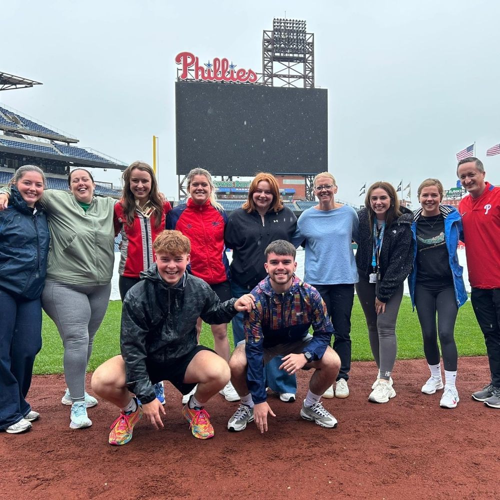 A group of ten adults, including two males crouching in the foreground, stand on a dirt baseball field under a light rain. A large scoreboard with "Phillies" at the top is visible in the background, along with stadium seating and American flags.