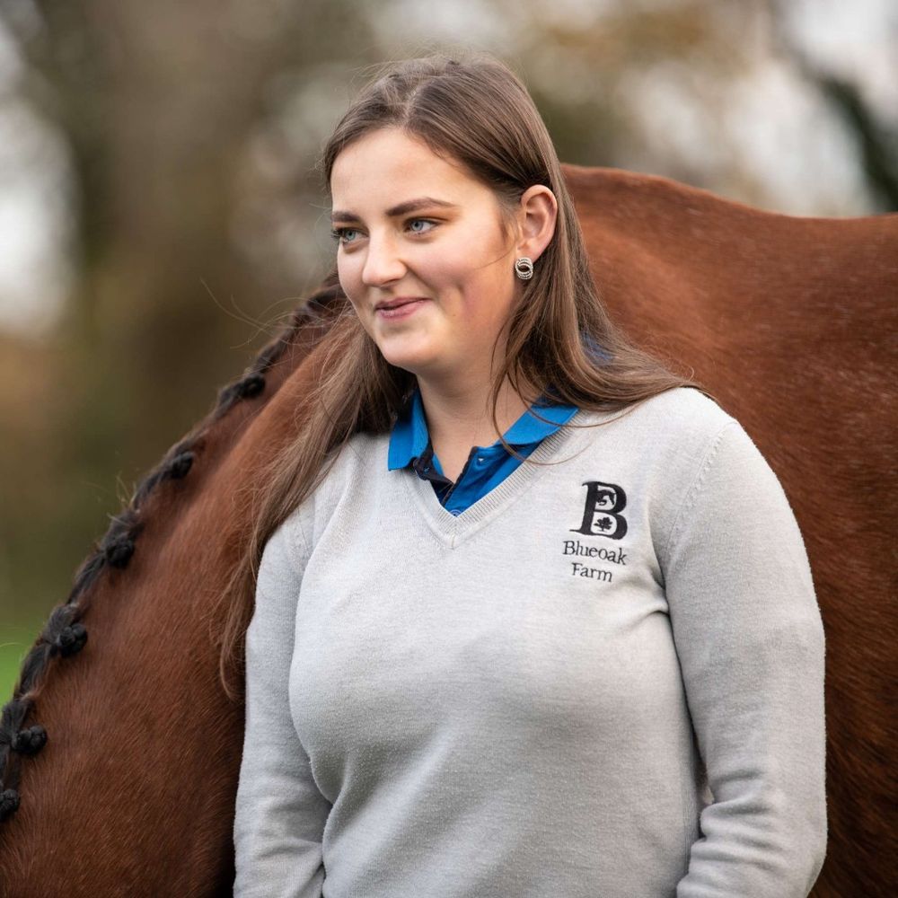 A young woman, Katherine Morton, wearing a gray v-neck sweater with the "Blueoak Farm" logo over a blue polo shirt, stands next to a brown horse whose mane is intricately braided.