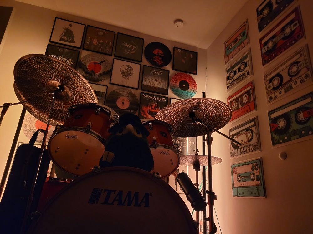 Corner with gallery walls of vinyl albums and decorative acoustic tiles. Lit up orange with Tama drum kit with Zildjian cymbals in the foreground. Plague doctor stuffed animal on top of the bass drum.
