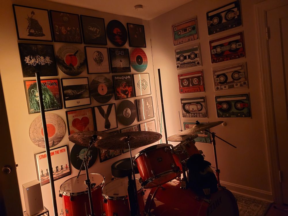 Corner with gallery walls of vinyl albums and decorative acoustic tiles. Lit up orange with Tama drum kit with Zildjian cymbals in the foreground. Plague doctor stuffed animal on top of the bass drum.