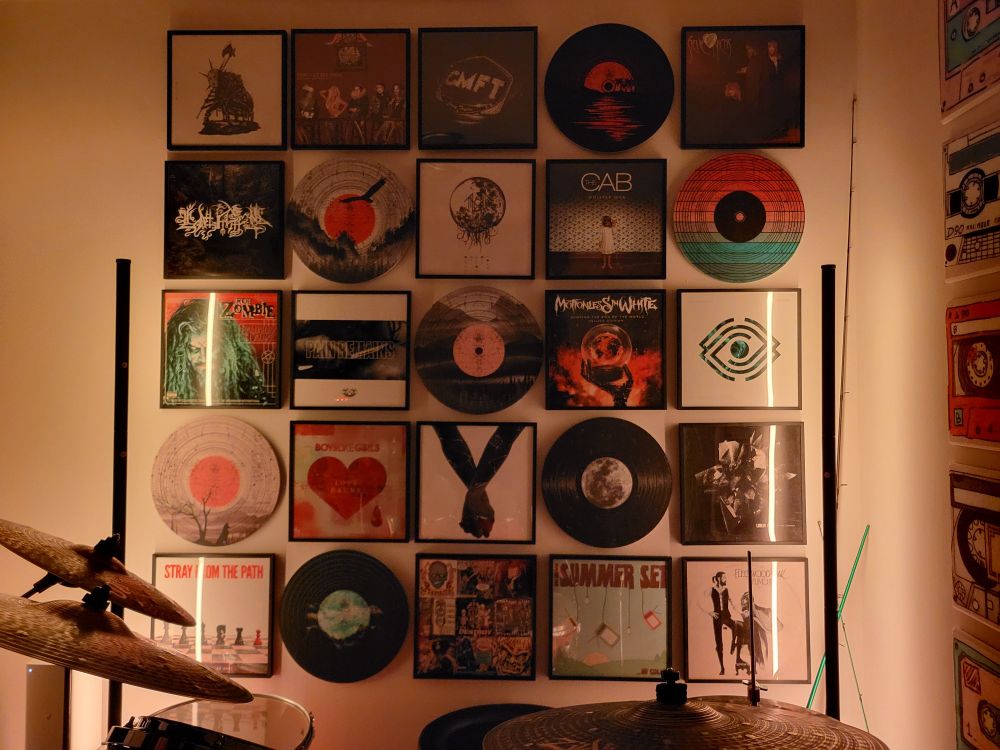 Gallery wall of vinyl albums and decorative acoustic tiles. Lit up orange. Albums: Finding God Before God Finds Me - Bad Omens, Only Death is Real - Stray From The Path, Take Me Back to Eden - Sleep Token, Pain Remains - Lorna Shore, Eternal Blue - Spiritbox, Scoring The End of the World - Motionless in White, ...In Color - The Summer Set, Whisper War - The Cab, Love Drunk - Boys Like Girls, A Fever You Can't Sweat Out - Panic! At The Disco, CMFT - Corey Taylor, Living Things - Linkin Park, Rumours - Fleetwood Mac, Hellbilly Deluxe - Rob Zombie, The Wild Heart - Stevie Nicks