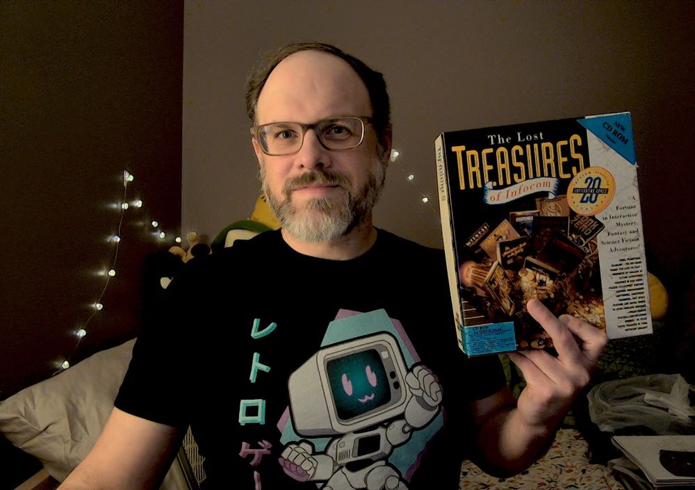 Photo of me wearing a DOSember shirt. It has an illustration of an adorable mascot that’s an anthropomorphized PC making an uwu face. On the side is some Japanese katakana that roughtly translates to “retro game.” I’m holding up a boxed copy of The Lost Treasures of Infocom.