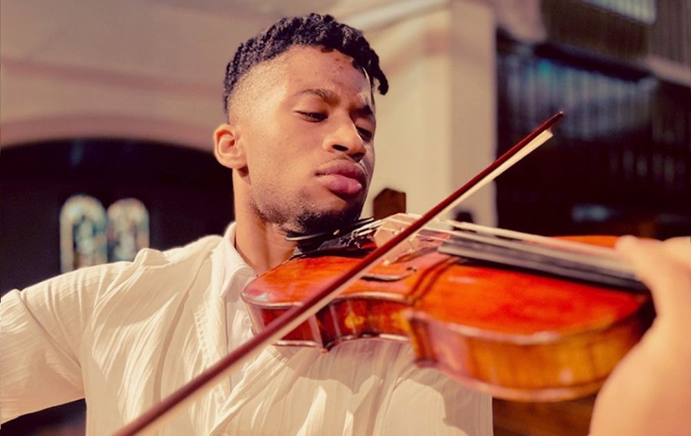 ​Violinist​ Irain Quirós, dressed in a textured white shirt, plays with intense focus under warm, dramatic stage lighting. The golden spotlights create a theatrical atmosphere, highlighting the rich wood tones of the violin and casting soft shadows around him. The background, a dimly lit concert hall with stained glass windows and organ pipes, adds depth and elegance to the scene.