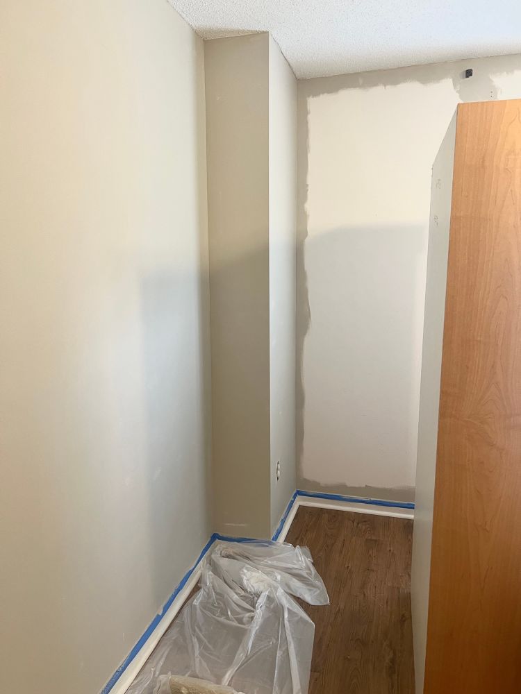 A corner in our guest room with some of the wall in sculptor clay and some white. There’s a plastic drop cloth on the floor and blue tape on the baseboard. 