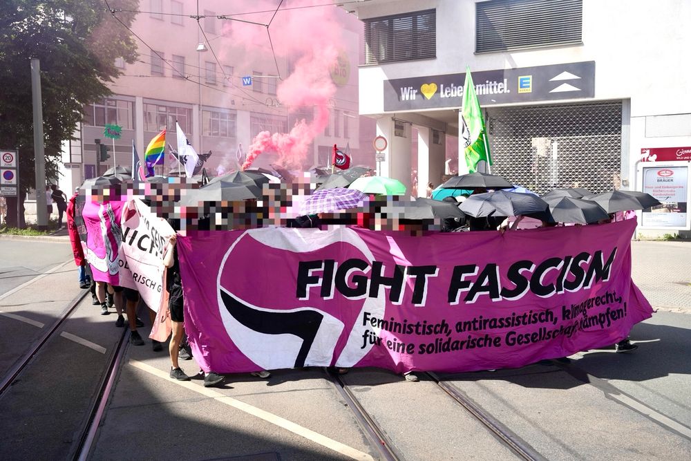 Auf dem Fronttransparent steht „Fight Fascism“ daneben ist ein Antifa Logo aufgemalt. Dahinter sind zahlreiche Regenschirme aufgespannt, ein Rauchtopf wurde gezündet 
