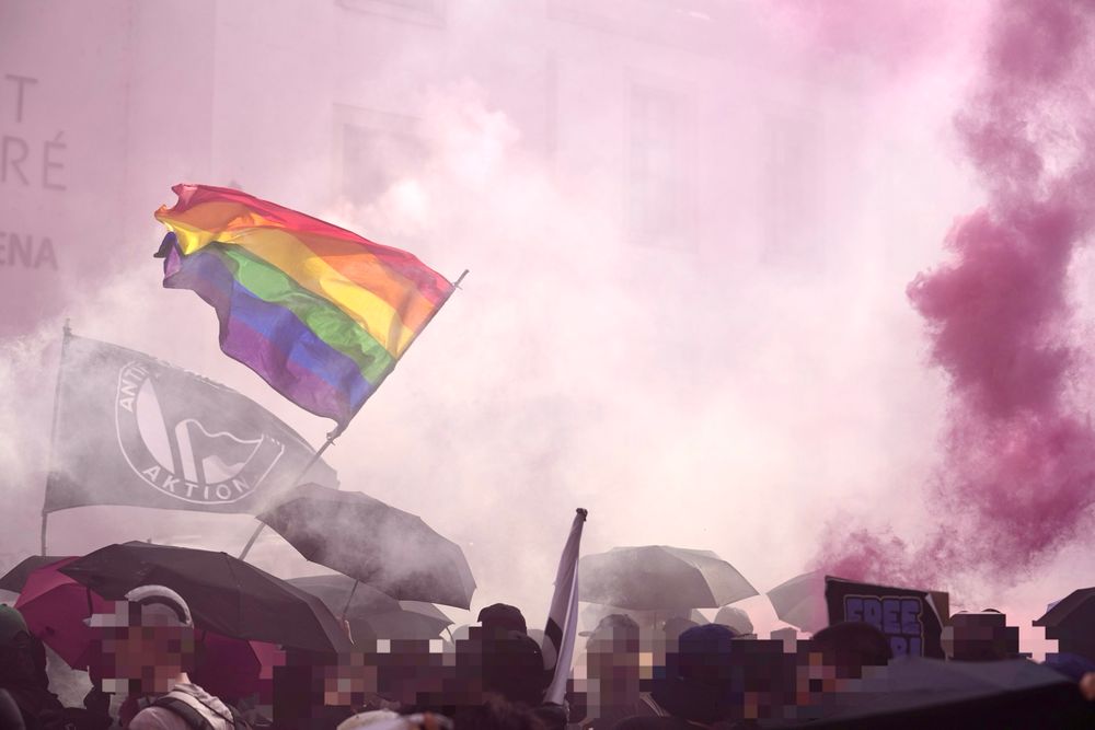 Eine Regenbogen-Fahne weht in rotem Rauch. Die Sonne scheint durch den Rauch auf die Demonstrant*innen, die Regenschirme tragen 