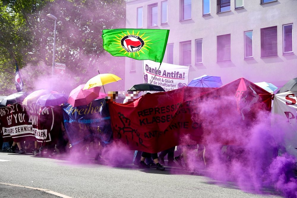 Lila Rauch steigt von einem Auf dem Boden liegenden Rauchtopf auf und hüllt die Demonstration in den Rauch. Eine grüne Antifa Fahne wird geweht. Die Teilnehmer*innen schirmen sich nach außen mit Regenschirmen ab.