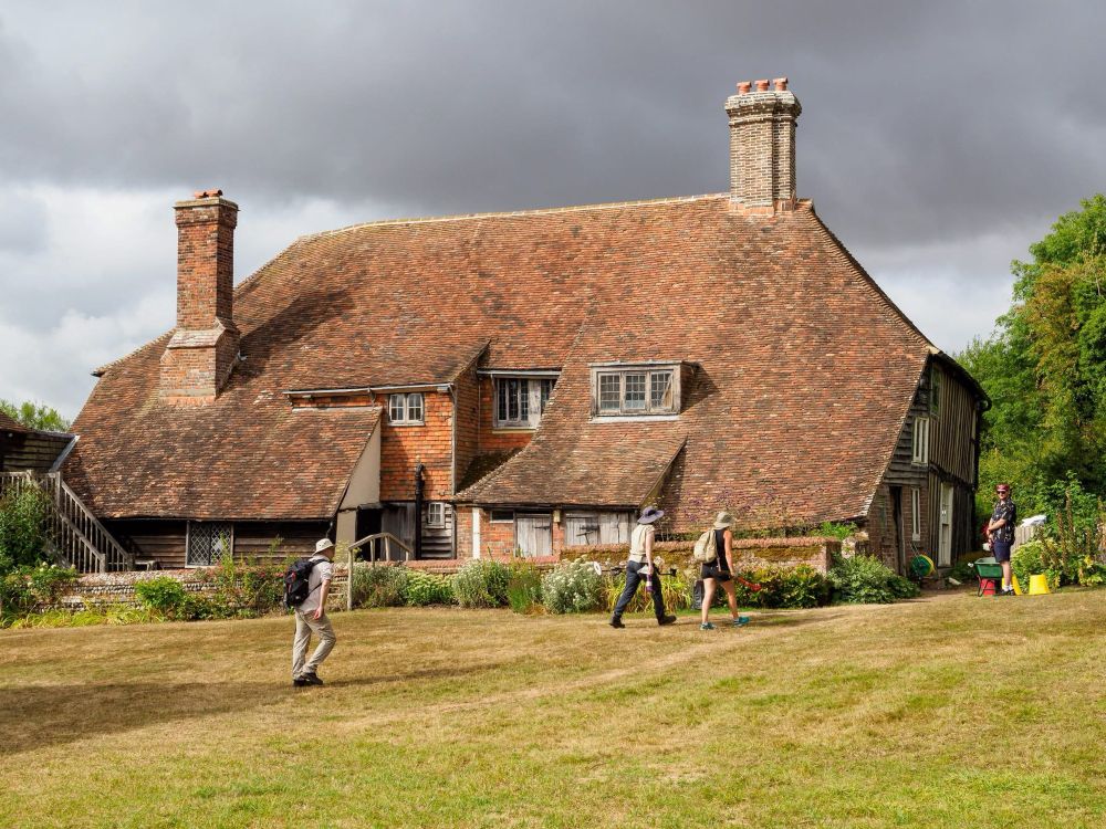 View of a late medieval house with a steeply sloping roof (a ‘cat slide’ roof) with a flower garden and lawns in front of it. A small group of people carrying equipment are crossing the garden. 