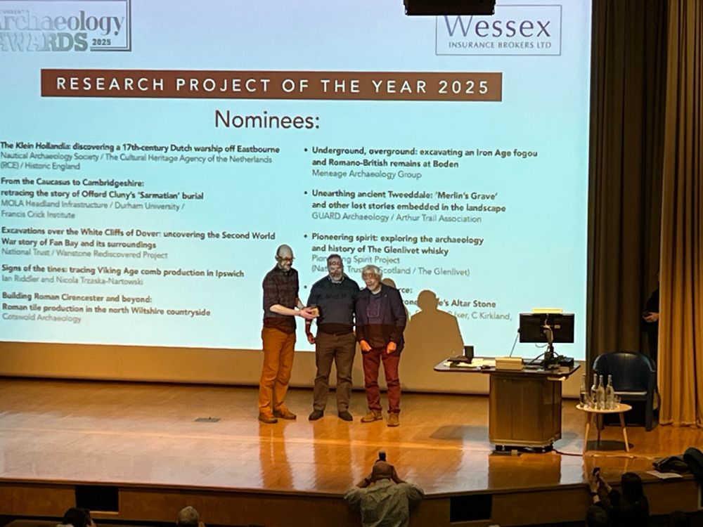 Three people on a large stage. Archaeologists from National Trust Scotland receiving an award during the ceremony at Current Archaeology Live Conference.