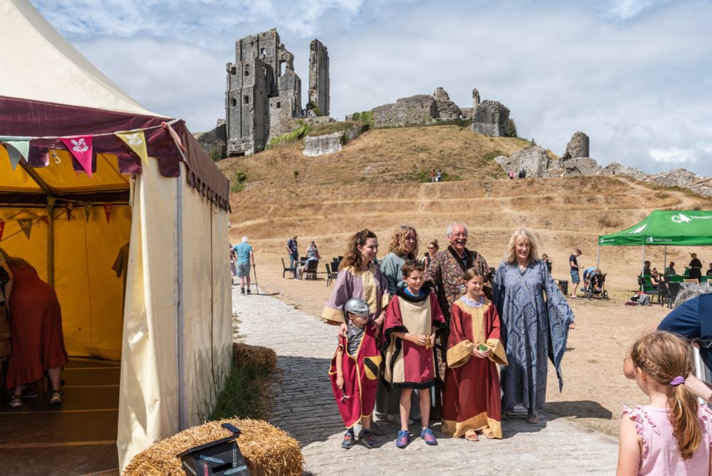 A group of people dressed up in medieval clothing stood next to a tent with the ruins of Corfe Castle on the hill behind
