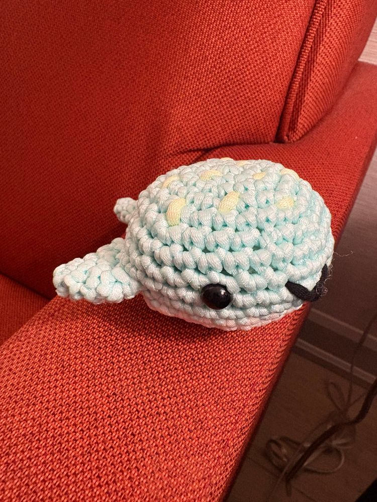 Light blue crocheted sting ray. The ray has black eyes, a black smile, and yellow spots on the head. You can see one fin and the tip of the tail. The bottom is white. The sting ray is on the arm of a rust orange couch.