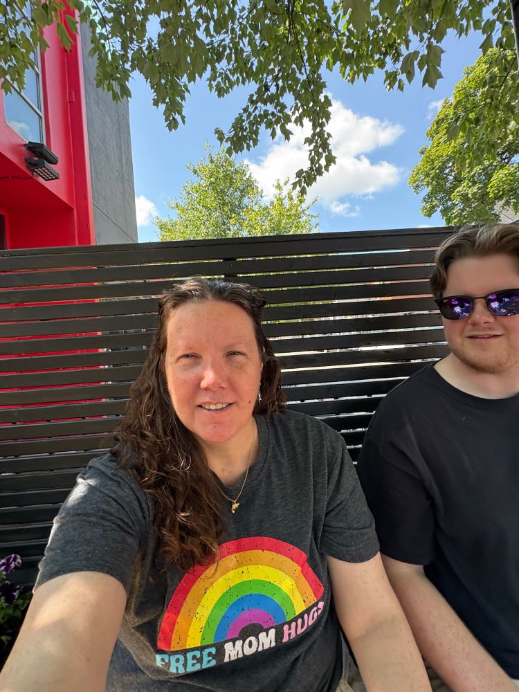 Me, sitting in the shade, sunglasses on my head, wearing a tshirt with a rainbow with the words “Free Mom Hugs”. And for the record, I mean that. One of my adorable & amazing kids is sitting beside me with his sunglasses on.