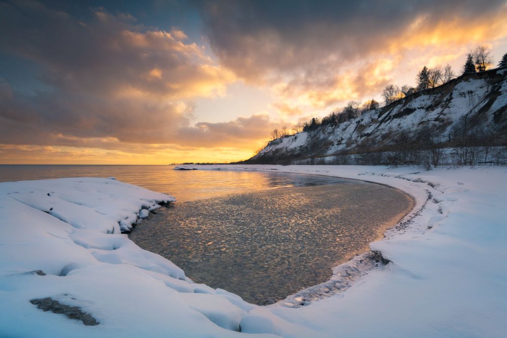 Snow-covered shoreline at sunset with a partially frozen body of water reflecting golden hues. A steep, tree-lined hill in the background is dusted with snow, contrasting against the dramatic sky filled with warm and cool tones. The scene captures the peaceful transition of day to night in a winter landscape.