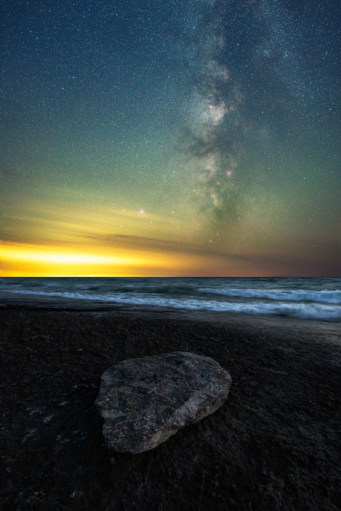 A serene nighttime coastal scene with the Milky Way galaxy stretching vertically across the star-filled sky. The horizon glows with warm hues of yellow and orange, reflecting off the gentle waves of the ocean. In the foreground, a large rock rests on the dark, textured shoreline.