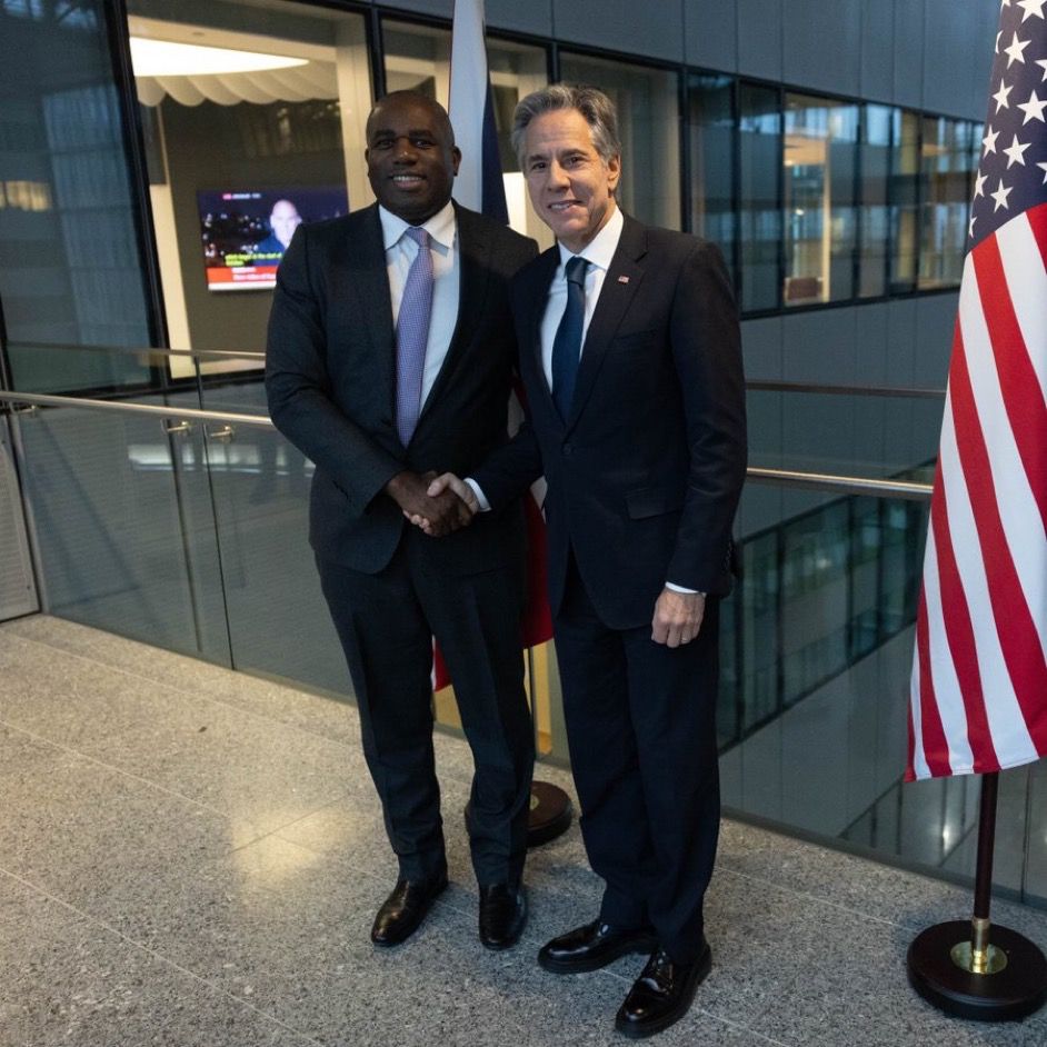 Secretary Blinken and UK Foreign Secretary Lammy shake hands and face forward smiling in Brussels on November 13, 2024. A U.S. flag is displayed to their left. (State Department/Chuck Kennedy)