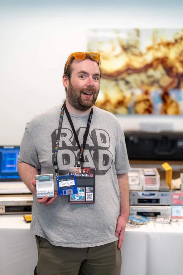 portrait photo of me standing awkwardly making a weird smile, with my gray rad dad shirt, a VCFSW blue (session presenter) badge as well as an MDCon Minidisc Mafia badge on, holding a silver MZ-B100 minidisc recorder, standing in front of my display, which is a bit bokeh'd out, I also have orange sunglasses propped up at my hairline, and I have some clear signs of sunburning because I kept forgetting big hats and long sleeves