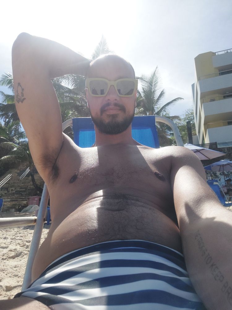 selfie sentado em uma cadeira reclinável na praia, com o braço direito levantado com a mão apoiada atrás da minha cabeça. estou vestindo uma sunga branca com listras pretas e óculos escuros com aro verde claro.