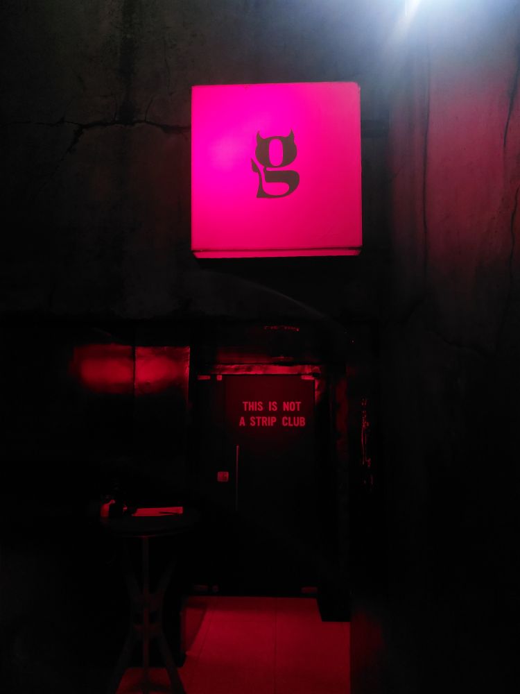 entrada escura de um bar com a letra G cursiva fazendo alusão ao diabo (com orelhas pontiagudas acima e um rabinho do lado direito) em um quadro iluminado de vermelho. na parede ao fundo, há a seguinte frase estampada: this is not a strip club.