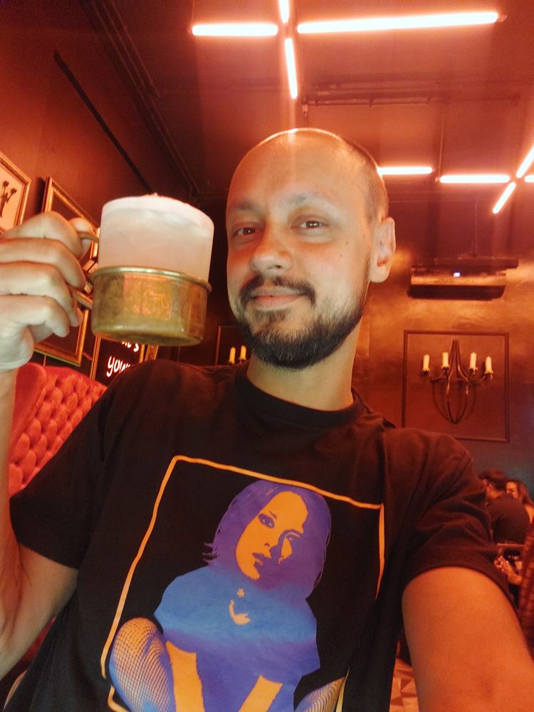 selfie segurando um drink de moscow mule em uma caneca prateada com a mão direita. estou vesindo uma camiseta preta com a cantora shirley manson estampada no peito em cores azul e laranja. ao fundo, há um candelabro preso na parede e linhas de luzes neon se ceuxando no teto com uma luz avermelhada no ambiente.