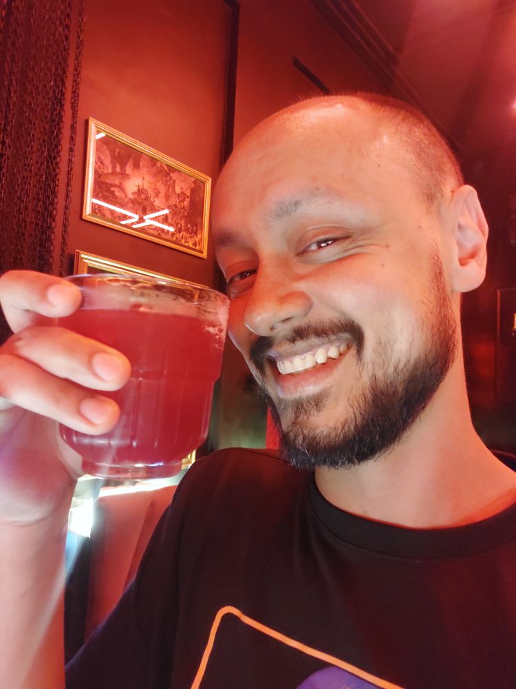 selfie segurando um drink misterioso de cor avermelhada com a mão direita. estou sorrindo e com olhos semi-cerrados  e vestindo uma camiseta preta.