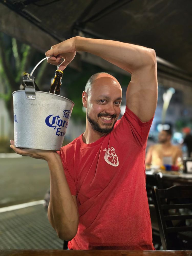 foto minha sentado em um bar, na calçada, segurando um balde com 3 garrafas de cerveja dentro (com uma só à mostra) com as duas mãos, deixando o braço direito arqueado por cima da cabeça. estou vestindo uma camiseta vermelha com o desenho de um coração humano em branco no peito esquerdo. 