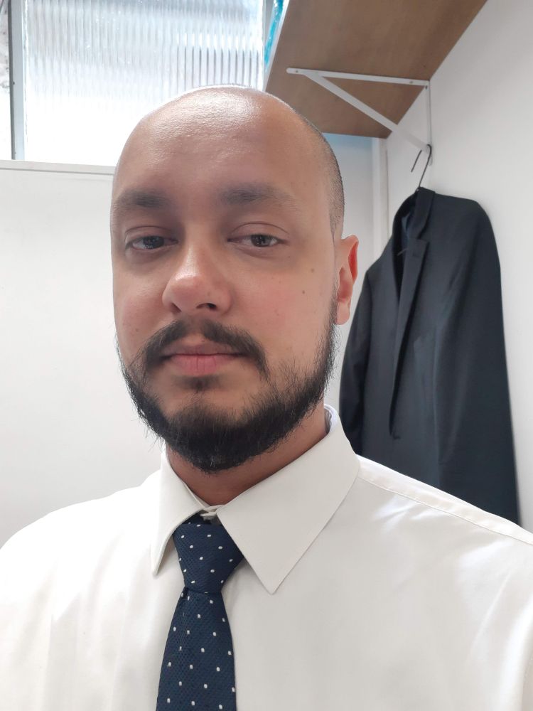 selfie vestindo camisa branca e gravata azul marinho com bolinhas brancas. atrás está um tento cinza escuro pendurado em um cabide debaixo de uma prateleira de madeira, com um vitrô ao lado. 