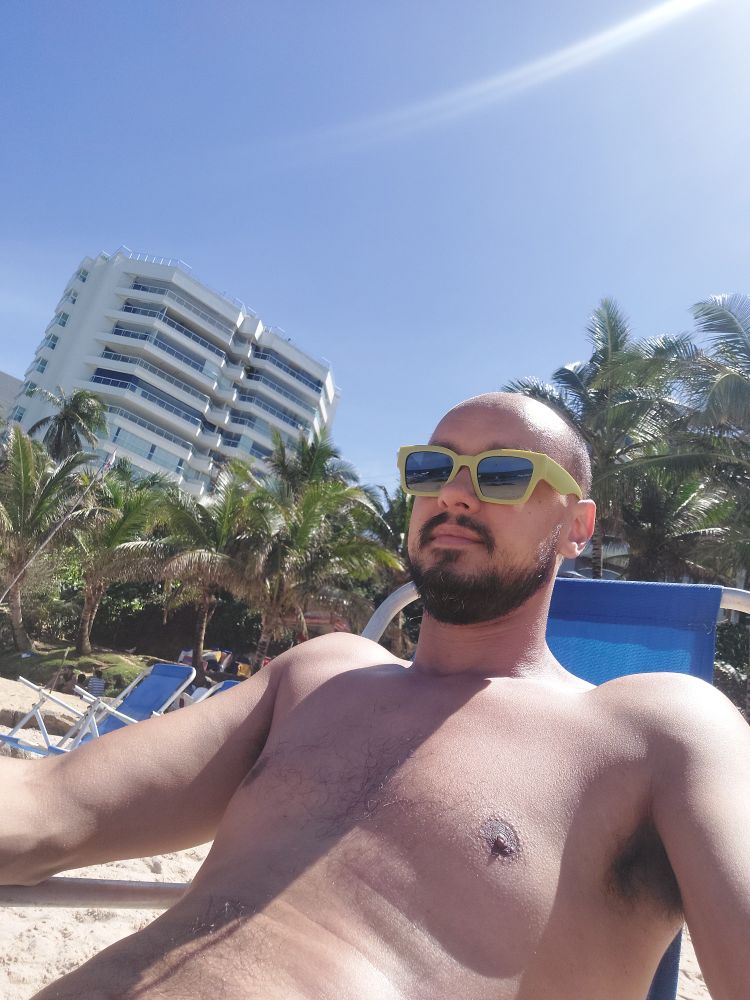 selfie sentado em uma cadeira reclinável na praia, sem camiseta e de óculos escuros de aro verde claro, com um prédio ao fundo.