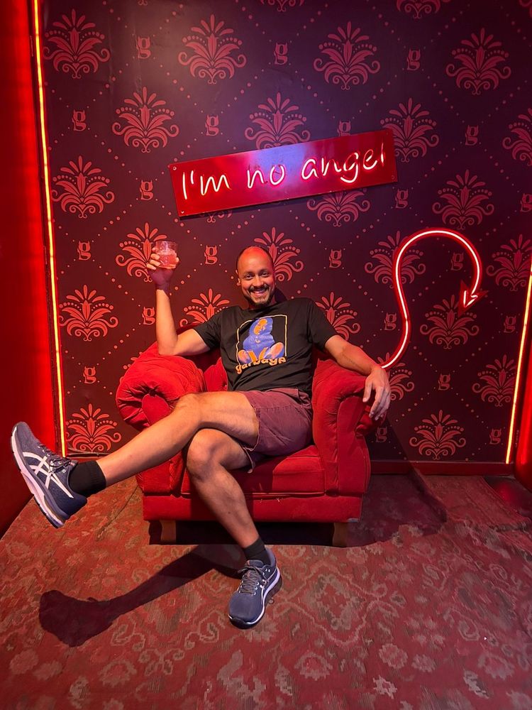 foto sentado em um sofá vermelho, vestindo camiseta preta e bermuda vinhoz meia preta e tênis azul. na parede, há uma placa com a escrita "i'm no angel" em neon. na parede, há um desenho de um rabo de diabo em neon vermelho saindo atrás do sofá.