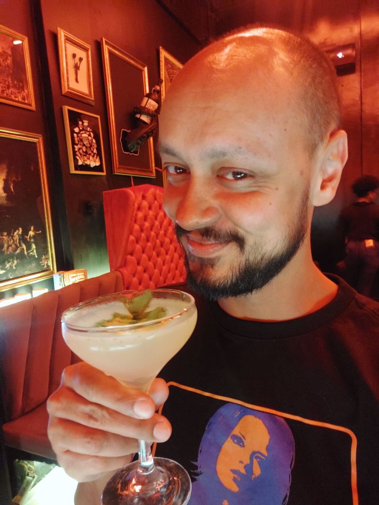 selfie segurando um drink que agora não sei o nome (é crítico) com a mão direita e com um sorrisinho maroto. ao fundo, quadros pendurados na parede e uma poltrona alta de cor vermelha. estou vestindo uma camiseta preta com a cantora shirley manson estampada no peito.