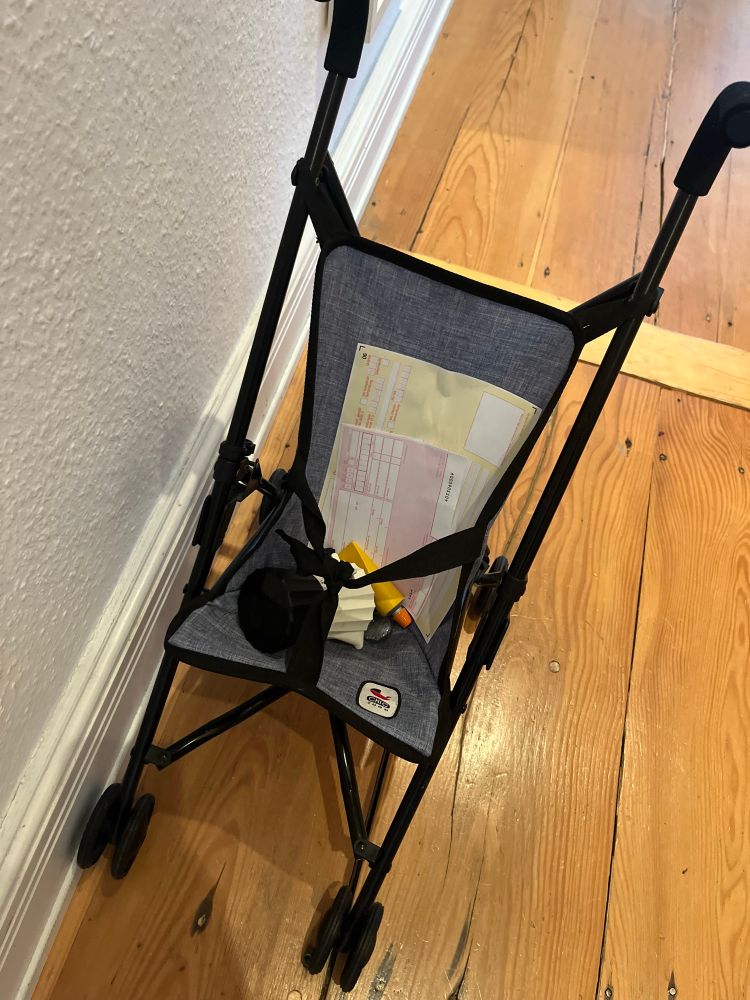 Man sieht einen von oben fotografierten Buggy für Puppen, in dem ein Rezept und ein Überweisungsschein, eine Espressokanne zum spielen und eine Tube Salbe aus Holz liegen.