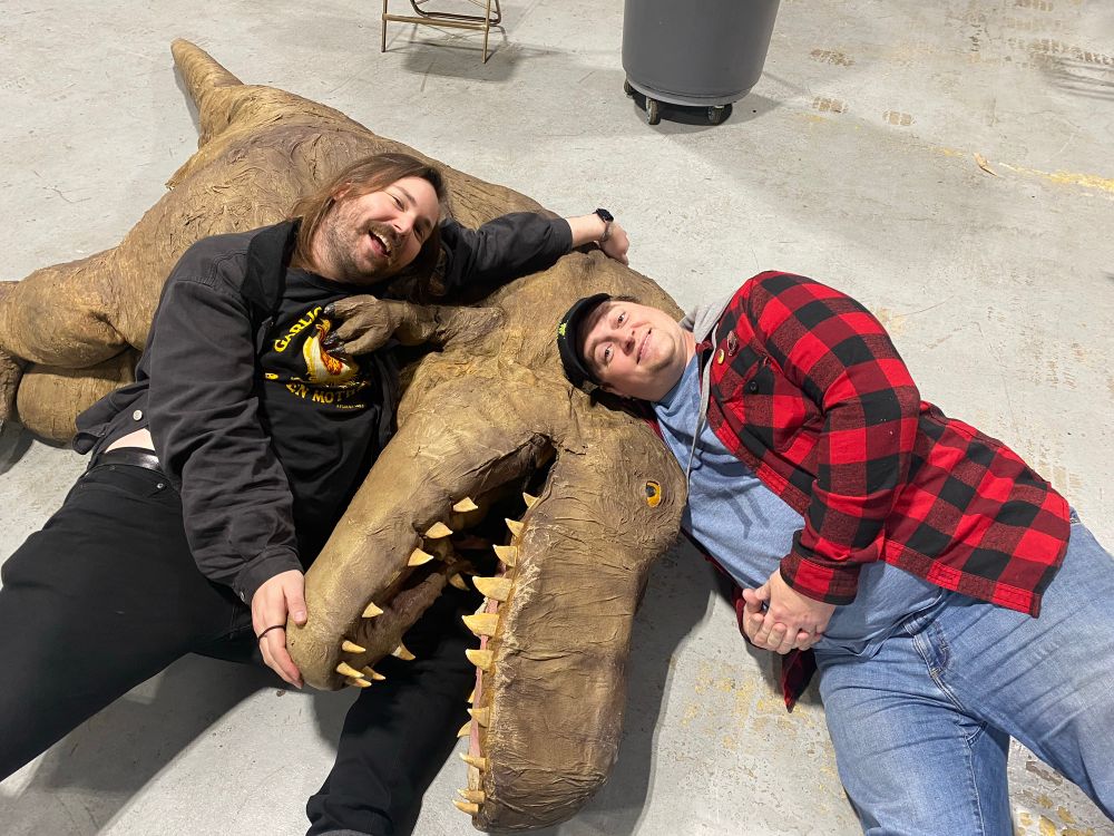Jesse Gouldsbury and Russ laying on the floor with Boone, the dinosaur from VelociPastor 