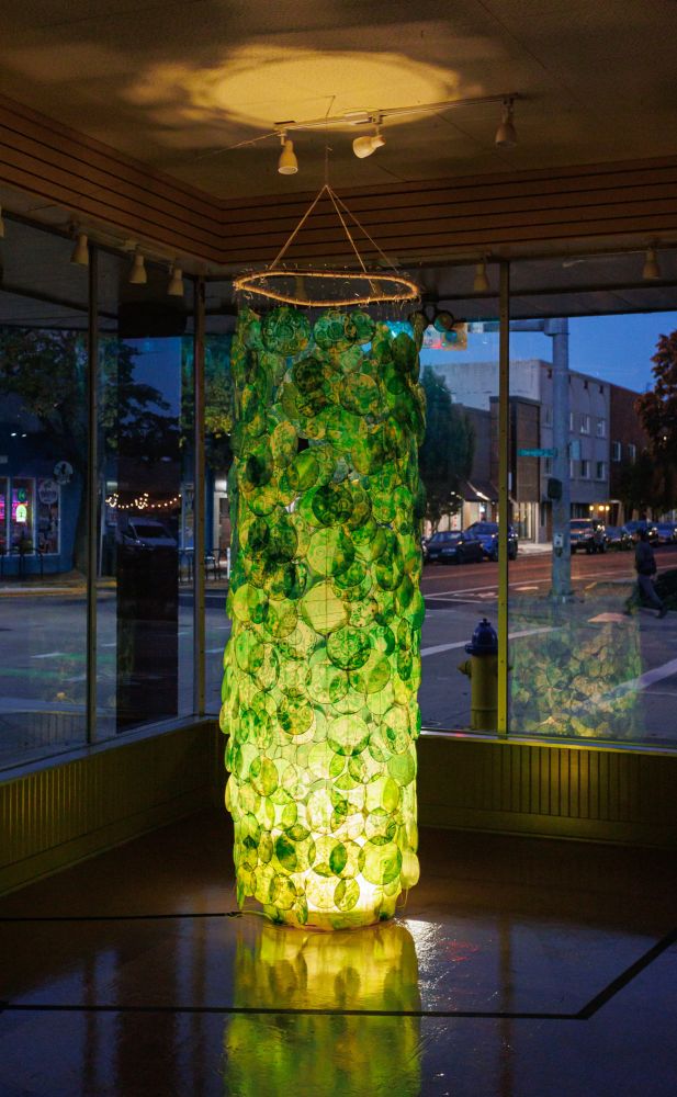 Installation art based on volvox algae, made of green paper circles forming a column, lit from below so it glows.