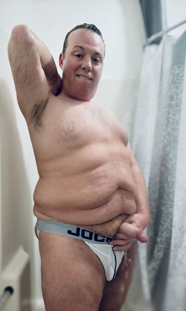 Tyler is posing in the shower with his fat cock out of his wet, white JockMail jockstrap. He has his hand behind his head showing off his hairy armpit and you can see water dripping off his cock. 