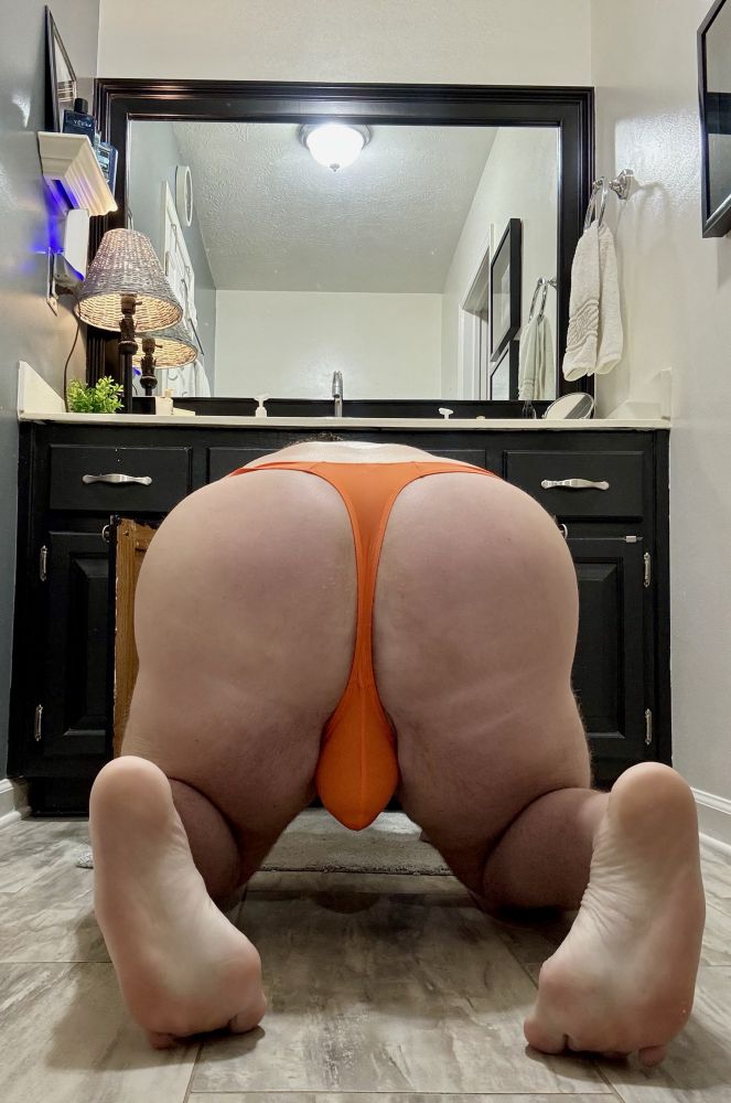 Tyler is on his hands and knees in the bathroom with his ass towards the camera, wearing an orange thong while searching in the bathroom cabinet for lube 