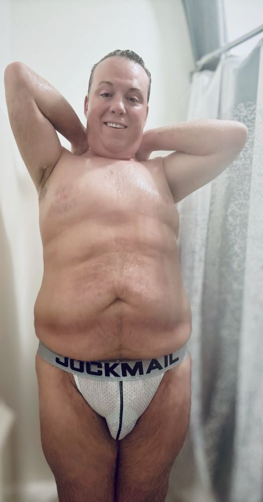 Tyler is posing in the shower wearing nothing but a JockMail white jockstrap that’s soaking wet. His hands are behind his head, exposing his armpits. 