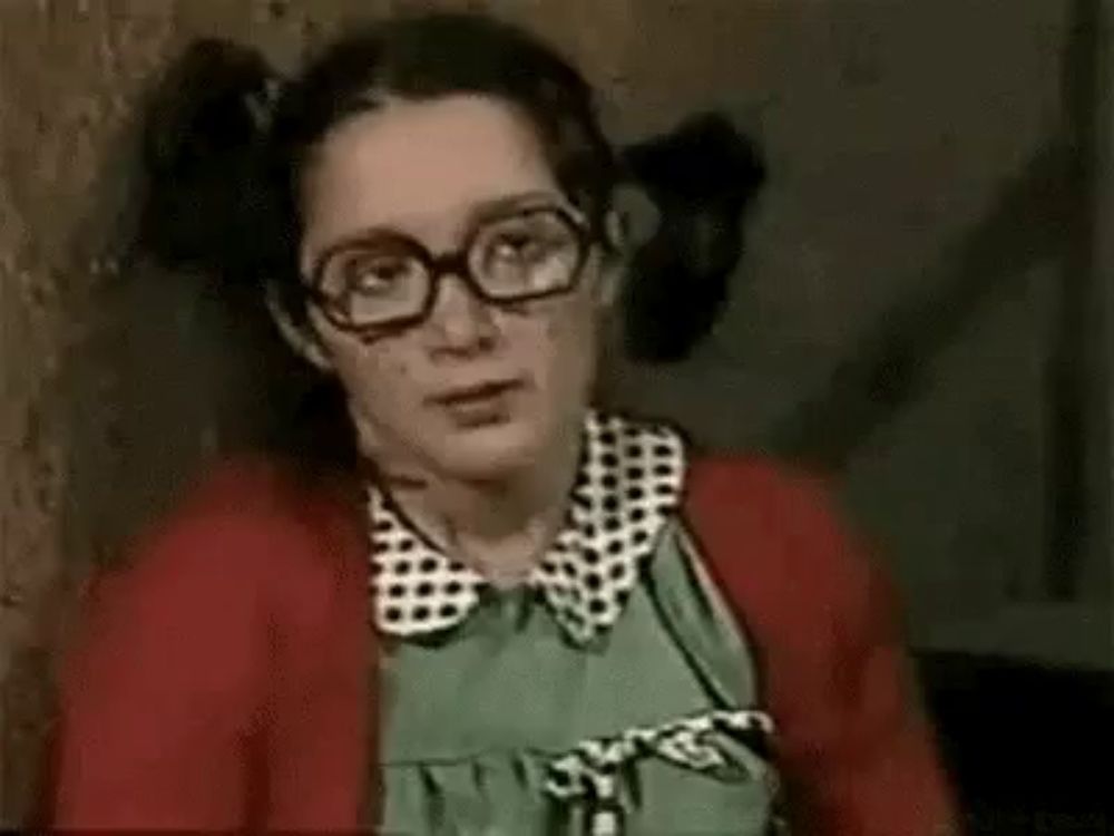 a woman wearing glasses and pigtails is leaning against a wall and making a funny face .