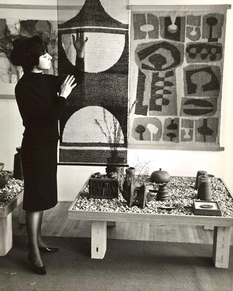 Akers examining one of her textiles, a long piece suspended in the middle of a room. Another textile piece is hanging on the wall behind her, and she is flanked by two low tables filled with pebbles displaying pieces of pottery.