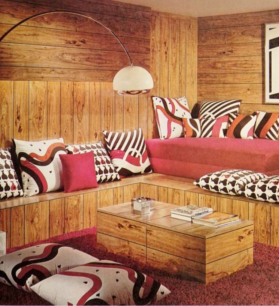A wood paneled lounge room with shag carpet and built in seating with pink blue black and white pillows with bold geometric patterns. From “The Young Decorator: a Textbook for High Schools and a Guide for all Home Decorators” by Phyllis Sloan Allen. 