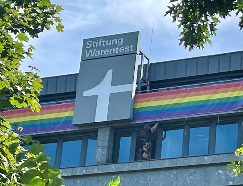 Ein Fassadenkletterer befestigt ganz oben am Gebäude der Stiftung Warentest ein langes Regenbogenbanner rechts und links des großen grau-weißen Stiftungslogos.