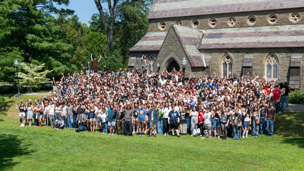 A large group of students pose for a group photo on a green lawn outside of a brick chapel.
