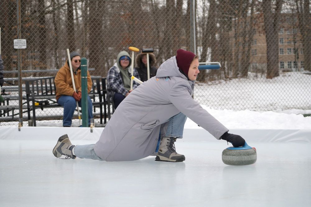 A woman in a gray winter coat slides a curling stone in front of her on the ice.