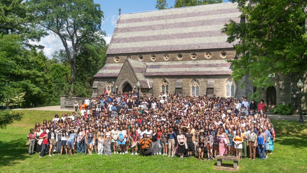 A group of Bard students pose for a photo in front of a stone chapel with lush green trees and grass all around them.