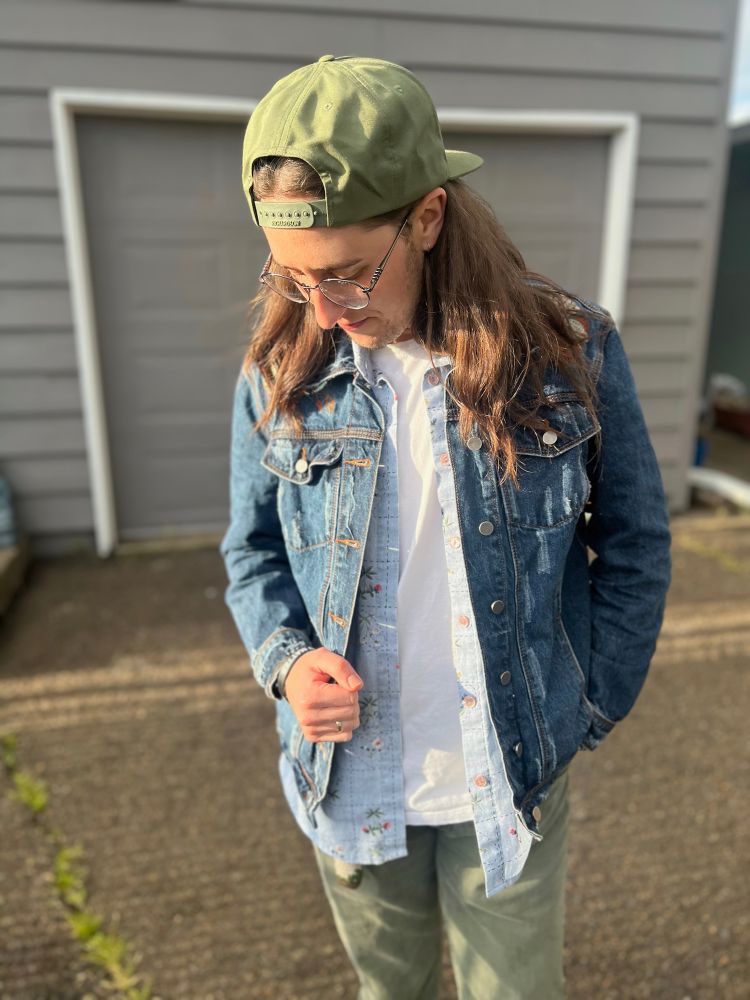 Quinn is looking down and standing in the driveway of their new duplex. They are wearing sage green jeans, a white t-shirt, a light blue button-up, a dark blue jean jacket, and a green snap-back. They have long brown hair and are lit up by the golden hour sunshine. 