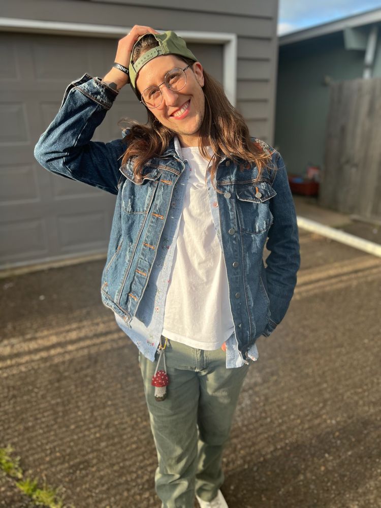 Quinn is smiling and standing in the driveway of their new duplex. They are wearing sage green jeans, a white t-shirt, a light blue button-up, a dark blue jean jacket, and a green snap-back. They have their right hand resting on top of their head. They have long brown hair and are lit up by the golden hour sunshine. 