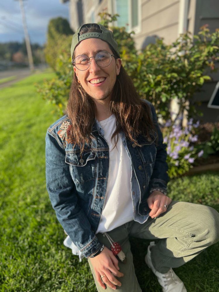 Quinn sits on a knee in front of their new duplex’s rose bush. They are smiling and wearing sage green jeans, a white t-shirt, a dark blue jean jacket, and a backwards green snap-back. They have long brown hair and are lit up by the golden hour sunshine. 