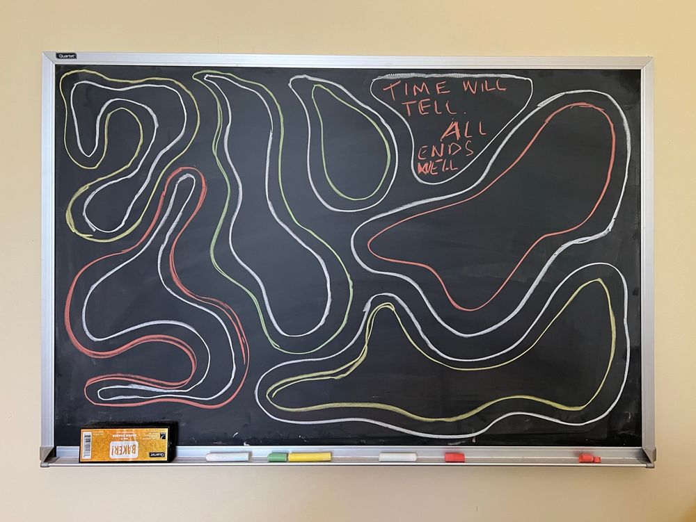 Multicolored wandering lines drawn like paths on a blackboard hung on a light yellow wall. At the top, someone has written “Time will tell. All ends well” in one of the sinuous spaces between the snaking lines. 