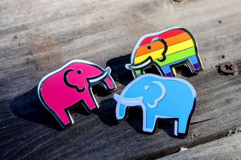 3 pins sit on a wooden table, one is a pink elephant, one is a blue elephant, and one is a rainbow elephant. They are very shiny and happy to be here.