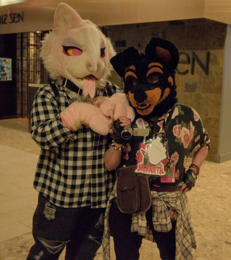 Fursuit photo of 2 people. Representing Kobra, a pink demon dog and Kenneth, a black and brown dog