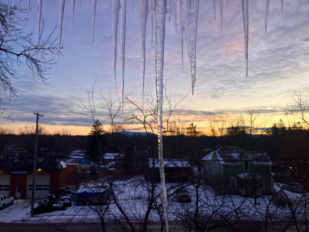 View of a snowy street at sunrise, with houses in foreground and mountains / trees in background, with a huge icicle running straight through the center of the view 